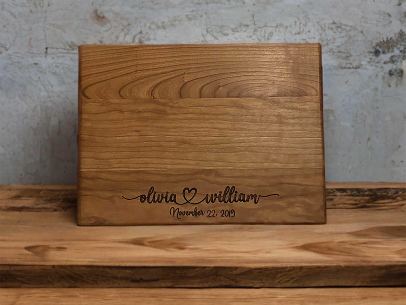 The image depicts a personalized wooden cutting board with engraving. The engraving includes two names with a heart symbol between them, followed by the anniversary date. The board is made of natural wood, showing visible grain patterns