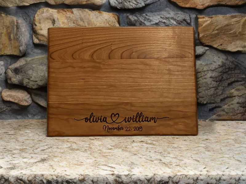 The image depicts a personalized wooden cutting board with engraving. The engraving includes two names with a heart symbol between them, followed by the anniversary date. The board is made of natural wood, showing visible grain patterns