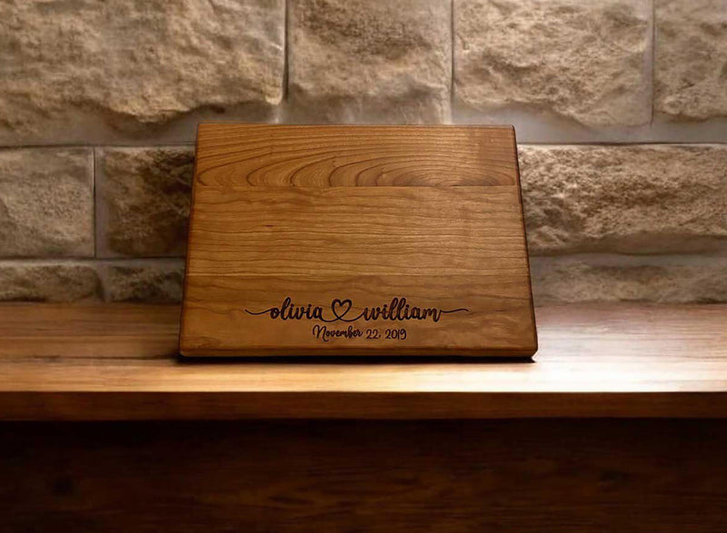 The image depicts a personalized wooden cutting board with engraving. The engraving includes two names with a heart symbol between them, followed by the anniversary date. The board is made of natural wood, showing visible grain patterns