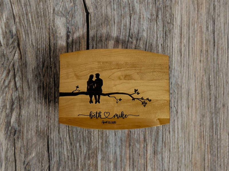 A wooden board with an engraving of a couple sitting on a tree branch, surrounded by small blossoms. Below, personalized names and a date are engraved in elegant script. The natural wood grain contrasts with a rustic wooden backdrop.