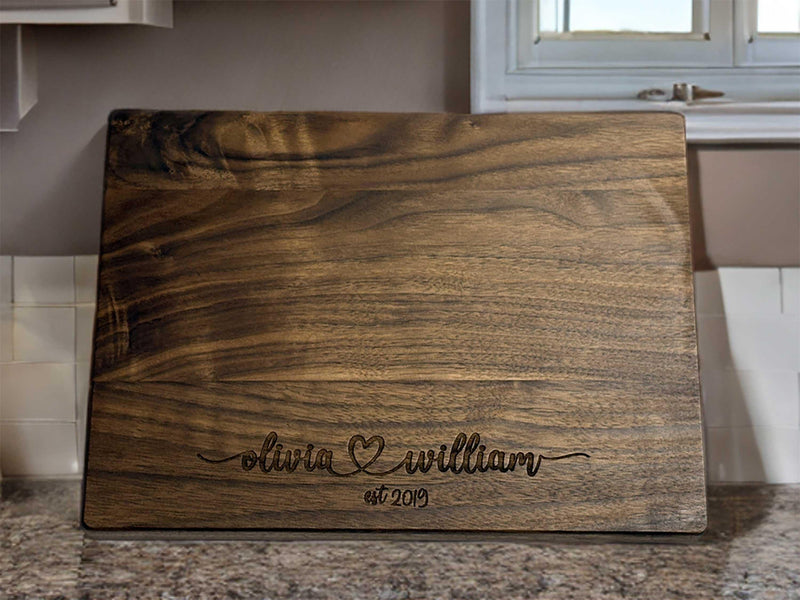 The image depicts a personalized wooden cutting board with engraving. The engraving includes two names with a heart symbol between them, followed by the anniversary date. The board is made of natural wood, showing visible grain patterns