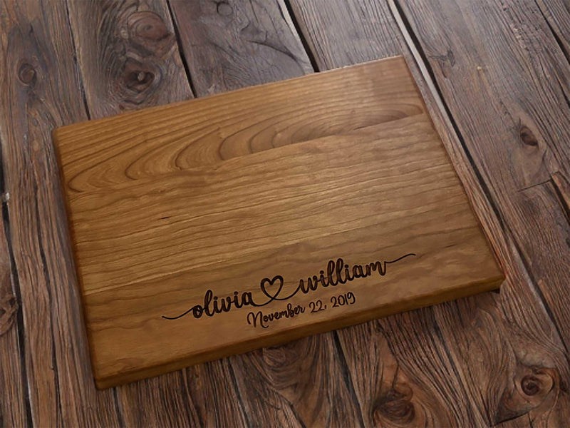 The image depicts a personalized wooden cutting board with engraving. The engraving includes two names with a heart symbol between them, followed by the anniversary date. The board is made of natural wood, showing visible grain patterns