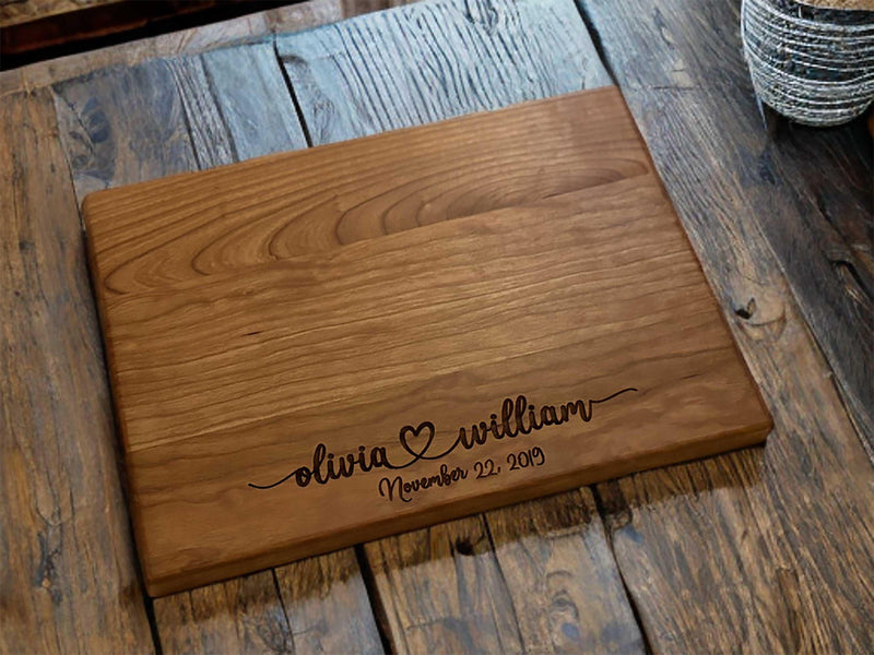 The image depicts a personalized wooden cutting board with engraving. The engraving includes two names with a heart symbol between them, followed by the anniversary date. The board is made of natural wood, showing visible grain patterns