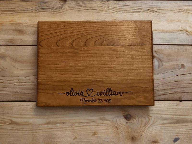 The image depicts a personalized wooden cutting board with engraving. The engraving includes two names with a heart symbol between them, followed by the anniversary date. The board is made of natural wood, showing visible grain patterns