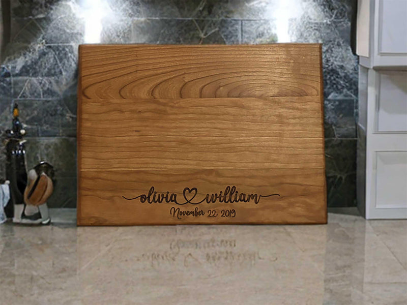 The image depicts a personalized wooden cutting board with engraving. The engraving includes two names with a heart symbol between them, followed by the anniversary date. The board is made of natural wood, showing visible grain patterns